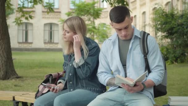 Charmantes blondes Mädchen, das einem jungen, gutaussehenden Jungen Kopfhörer gibt, der in ein Buch vertieft ist. Porträt von befreundeten Studenten, die an einem sonnigen Tag auf dem Campus-Hof sitzen. Paar rastet aus und lernt im Freien. — Stockvideo