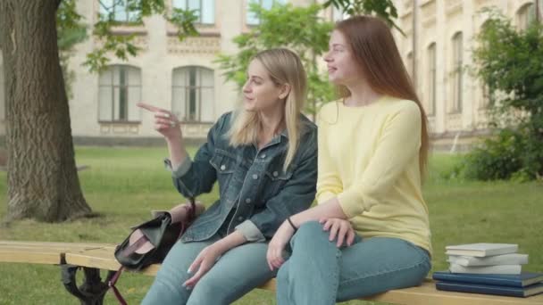 Dos alegres estudiantes caucásicas sentadas en el patio de la universidad y mirando hacia otro lado. Retrato de la joven hermosa rubia y pelirroja hablando al aire libre en el descanso. Estilo de vida, educación, ocio . — Vídeo de stock