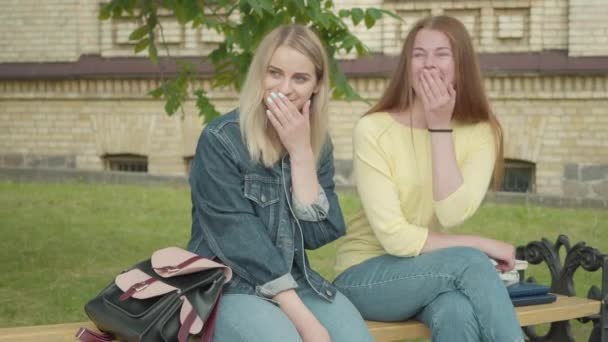 Pozitív vörös hajú lány suttog a barátainak, miközben az egyetemi udvaron ül a padon. Vidám, elbűvölő kaukázusi diákok csukott szájjal nevetnek hangosan. Pletykák, pletykás barátság.. — Stock videók