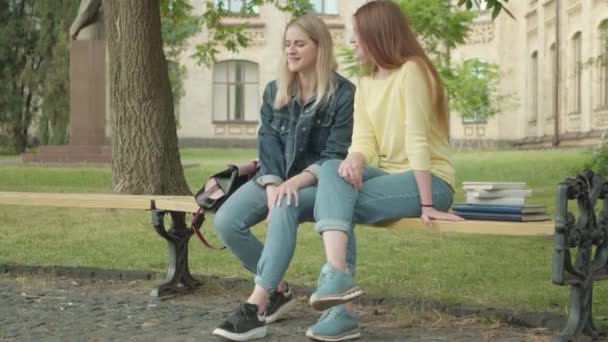 Bredbild av två flickor som sitter på bänken utomhus och pratar. Leende positiva kaukasiska studenter vilar på universitetets gård. Vänskap, utbildning, livsstil. — Stockvideo