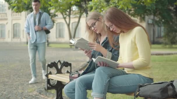 İnek beyaz kızlar genç yakışıklı sporcu geçerken utangaçlaşıyorlar. Gözlüklü güzel bayan öğrenciler kampüs bahçesinde kitap okuyorlar.. — Stok video