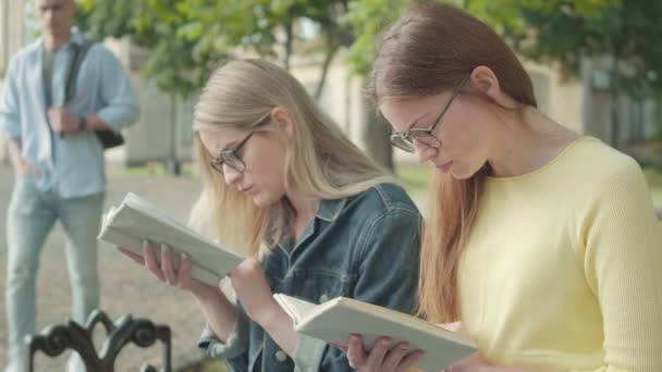 Close-up zijaanzicht van gefocuste meisjes in brillen die zich verschuilen achter boeken als knappe atletische sporter die langskomt. Portret van verlegen blonde en roodharige vrouwelijke studenten in de tuin van de universiteit. — Stockvideo
