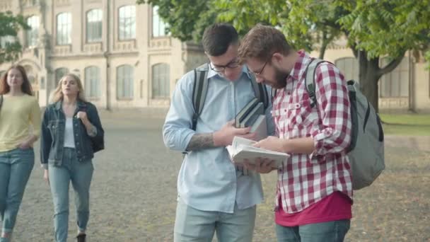 Nerds masculinos hablando como hermosas chicas modernas que pasan por aquí. Retrato de tímidos estudiantes inteligentes ajustando anteojos mirando a magníficos compañeros de grupo en el patio de la universidad. Coqueteo, educación, estilo de vida . — Vídeos de Stock