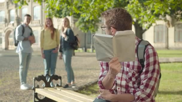 Tímido estudiante universitario caucásico en gafas que sufren de intimidación de compañeros de grupo. Retrato de un joven barbudo sentado en el banco con un libro mientras la gente borrosa se ríe del fondo . — Vídeos de Stock