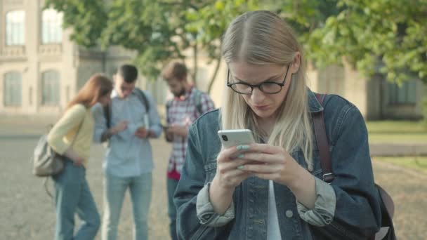 Blond meisje scrollen smartphone scherm als wazig groepgenoten lachen op de achtergrond. Nerd jonge blanke student in bril die lijdt aan online pesten op de universiteit. — Stockvideo