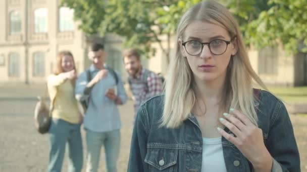 Portrét ztrapněné dívky v brýlích, jak se dívá do kamery, zatímco ji rozmazaní spoluhráči šikanují v pozadí. Plachý inteligentní dívka ignoruje výsměch na univerzitě. — Stock video