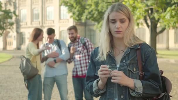 Die schöne schüchterne Blondine lächelt in die Kamera, während die Leute im Hintergrund über sie lachen. Selbstbewusste, intelligente kaukasische Studentin, die den Spott ihrer Mitschüler ignoriert. Intelligenz, Vertrauen. — Stockvideo