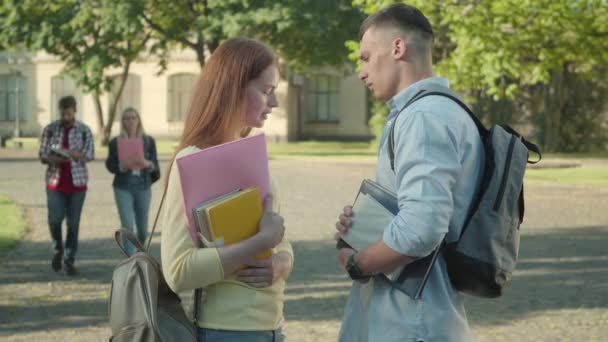 Vue latérale d'un jeune homme confiant qui demande à sortir avec une rousse sur le campus. Portrait d'étudiants caucasiens amoureux parlant en plein air lors d'une journée ensoleillée d'été. Amour, flirt, mode de vie. — Video