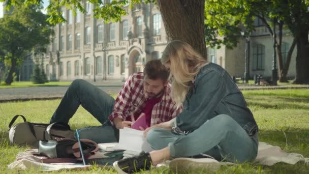 Szőke kaukázusi lány és barna szakállas fiú, akik könyveket és laptopot használnak. Portré modern egyetemi hallgatókról, akik a szabadban beszélgetnek a zöld réten. Oktatás, életmód, kommunikáció. — Stock videók