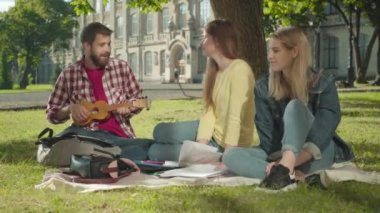 Üç neşeli öğrenci yeşil çimlerde otururken şarkı söylüyor ve gülümsüyor. Ukulele çalan beyaz adamın portresi kampüs bahçesinde dans eden neşeli kadınlar gibi. Üniversite yaşam tarzı, eğitim.
