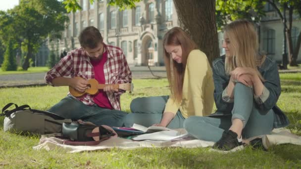 Hombre caucásico positivo jugando ukelele para chicas hermosas en el prado verde soleado. Retrato de jóvenes estudiantes de ambos sexos descansando en el patio del campus universitario. Ocio, relajación, estilo de vida universitario . — Vídeo de stock