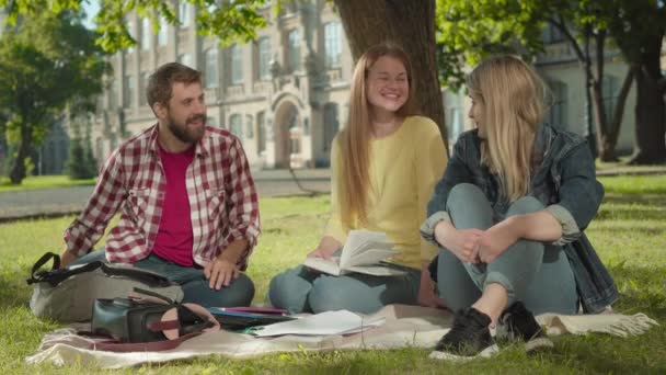 Tres alegres compañeros de grupo caucásicos hablando y riendo en el prado de verano en el patio del campus. Retrato de jóvenes positivos estudiando y descansando juntos en la universidad. Concepto de estilo de vida . — Vídeo de stock