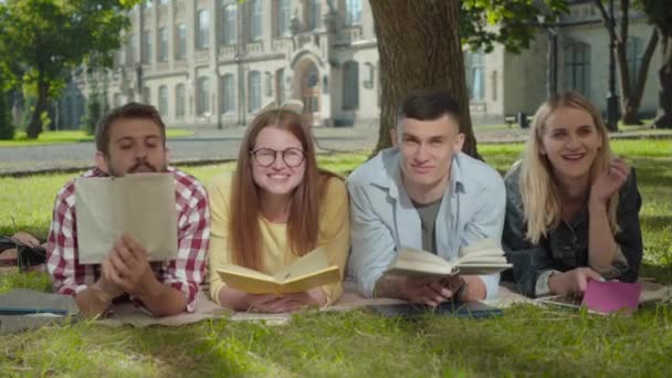 Retrato de felices estudiantes caucásicos, hombres y mujeres, acostados con libros sobre hierba verde y riendo. Cuatro hombres y mujeres alegres disfrutando descansando en el soleado patio del campus universitario. Estilo de vida . — Vídeos de Stock