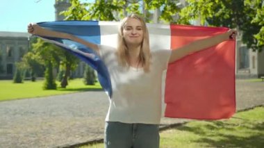 Elleri açık ulusal bayrağı tutan mutlu sarışın bir Fransız öğrencinin orta pozu. Yaz günü üniversite bahçesinde poz veren gülümseyen genç bir kadın. Vatanseverlik, eğitim, yaşam tarzı.