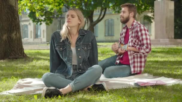 Portrait de jeune femme blanche blonde appréciant la musique comme homme barbu jouant de l'ukulélé en arrière-plan. Couple romantique d'étudiants assis sur la prairie verte sur la cour du campus universitaire. Amour, loisirs. — Video