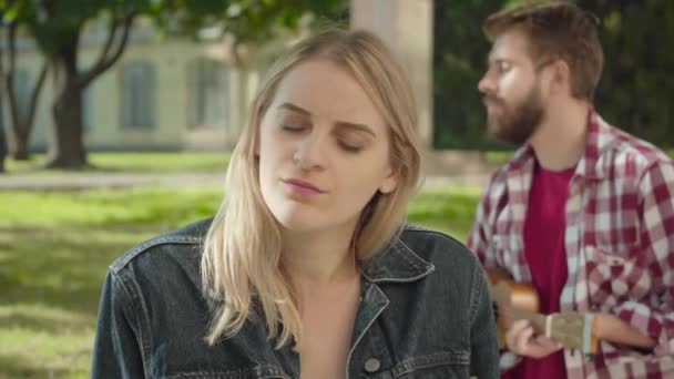 Cara de cerca de una joven rubia escuchando música y sonriendo. Hombre romántico borroso jugando ukelele en el fondo. Retrato de estudiantes universitarios caucásicos felices citas al aire libre . — Vídeo de stock