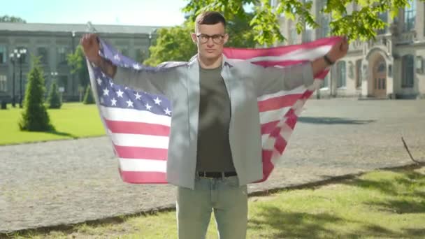 Joven confiado extendiendo las manos sosteniendo la bandera americana. Retrato de un guapo e inteligente estudiante caucásico posando en el patio de la universidad. Confianza, educación internacional, estilo de vida . — Vídeo de stock