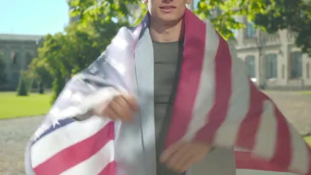 Felismerhetetlen amerikai diák, aki nemzeti zászlóba burkolózik. Egy fiatal férfi portréja, amint pózol az egyetemi udvaron a napsütéses napon. Életmód, hazafiasság, oktatási koncepció. — Stock videók