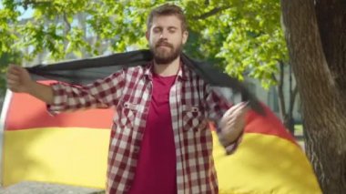 Kendini Alman bayrağına saran kendine güvenen sakallı üniversite öğrencisine kamera yaklaşıyor. Güneşli bir günde üniversite bahçesinde ulusal sembolle poz veren yakışıklı bir gencin portresi..