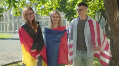 Bir grup uluslararası öğrenci üniversite kampüs bahçesinde sarılıp gülümsüyor. Güneşli yaz gününde açık havada poz veren Amerikan, Fransız ve Alman bayraklı kadın ve erkekler. Eğitim kavramı.