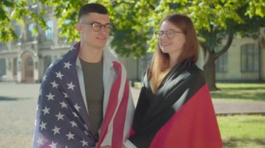 Üniversite kampüsünde poz veren mutlu Alman ve Amerikalı öğrencilerin portresi. Ulusal bayraklara sarılmış gözlüklü genç adam ve kadın kameraya bakıyor ve gülümsüyor..