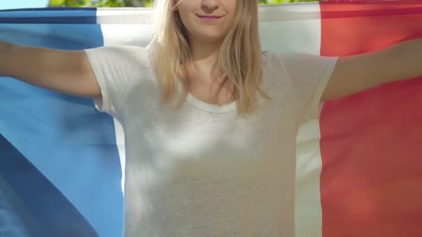 Eine bis zur Unkenntlichkeit lächelnde Frau hüllt sich an einem sonnigen Sommertag in eine französische Flagge. Porträt einer selbstbewussten, glücklichen Studentin, die mit Nationalsymbol auf dem Hof der Universität posiert. — Stockvideo