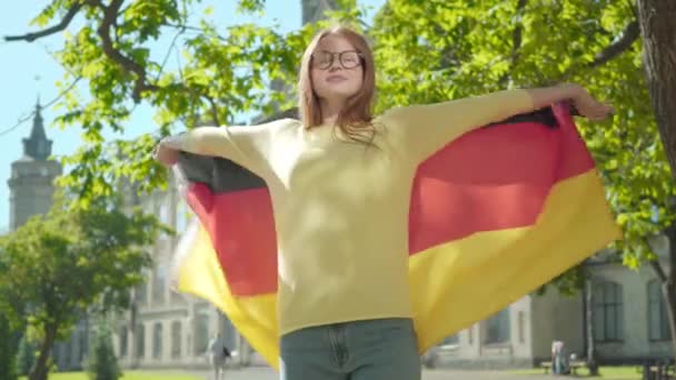 Vacker rödhårig kvinna i glasögon som sprider händer och snurrar med tysk flagga. Porträtt av ung vit kvinna student poserar i bakgrunden av universitetshuset på solig sommardag. — Stockvideo