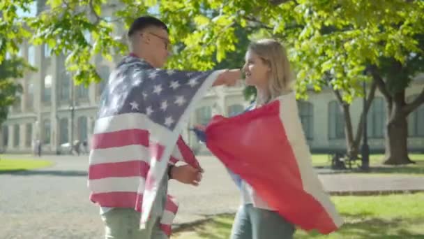 Sidovy av att älska internationella studenter kramas på soliga universitetsvarvet. Porträtt av positiv amerikansk man och fransk kvinna insvept i nationella flaggor. Utbildning, livsstil. — Stockvideo