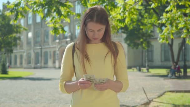 Mujer pelirroja joven contando dinero, poniendo dólares en el bolsillo trasero y cruzando las manos. Retrato de una encantadora estudiante caucásica sonriente posando en el patio de la universidad. Concepto independiente . — Vídeo de stock