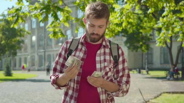 Portret pewnego siebie białego mężczyzny liczącego dolary na uniwersyteckim podwórku i wkładającego je do kieszeni. Młody przystojniak zarabiający pieniądze na studiach. Bogactwo, inteligencja. — Wideo stockowe
