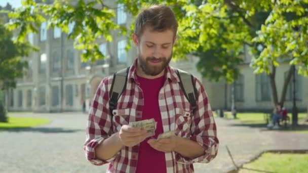 Boldog elégedett kaukázusi férfi számolja a pénzt az egyetem udvarán, és félrenéz. Portré fiatal férfi diák álmodik utazás napsütéses nyári napon. Életmód, vagyon, szabadúszó. — Stock videók