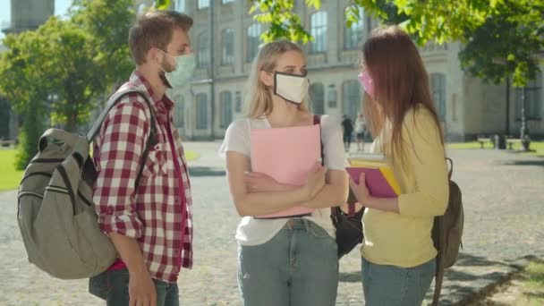 Drie studenten met gezichtsmasker praten op de zonnige campus. Portret van positieve blanke mannen en vrouwen die studeren aan de universiteit op Covid-19 pandemie. Coronavirusonderwijs en levensstijl. — Stockvideo