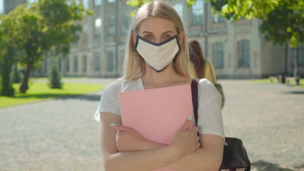 Kameraya bakan beyaz maskeli, sarışın, beyaz bir kadının portresi. Covid-19 karantinasında üniversite bahçesinde poz veren sırt çantası ve kağıtları olan bir kız öğrenci. Coronavirus pandemik yaşam tarzı. — Stok video