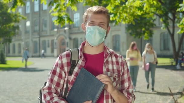 의료용 마스크를 입은 젊은 남성이 대학교 마당에 서서 카메라를 보고 지나가는 여성 학생으로 묘사되어 있습니다. Covid-19 전염병 격리 연구중인 백인 남자. — 비디오