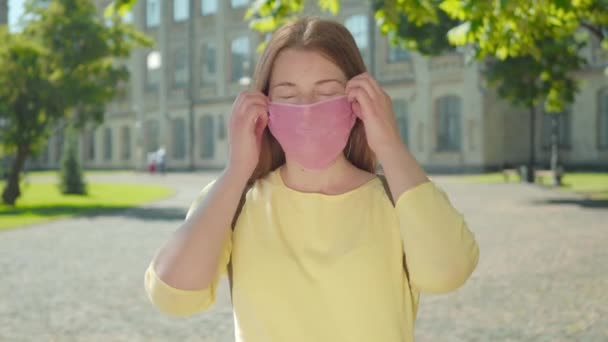 Femme rousse enlève le masque rose et respire. Portrait de belle étudiante caucasienne positive posant dans la cour de l'université sur le confinement pandémique Covid-19. — Video