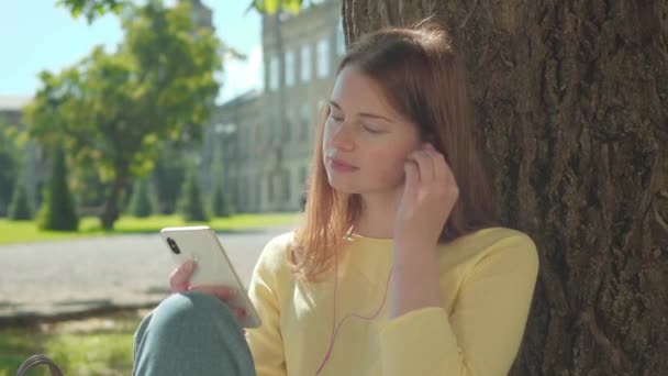 젊은 빨간 머리 여자가 이어폰을 끼고 스마트폰으로 음악을 켜고 있습니다. 아름다운 백인 여학생이 화창 한 여름날 대학교 운동장에서 휴식을 즐긴다. — 비디오