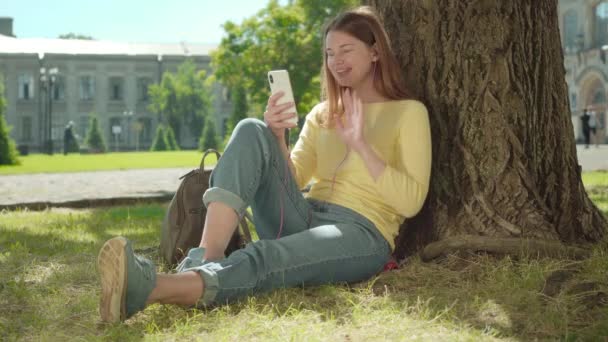 Chica pelirroja alegre utilizando el chat de vídeo al aire libre en el soleado día de verano. Retrato de una joven positiva caucásica sentada contra un árbol y hablando. Comunicación inalámbrica, estilo de vida, ocio . — Vídeos de Stock