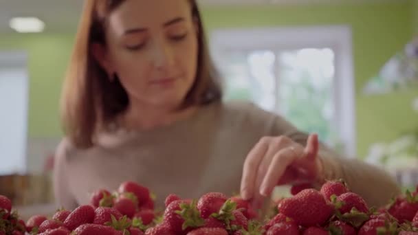 Primer plano de fresas rojas que yacen en el estante en el supermercado con una joven borrosa tocando y oliendo bayas en el fondo. Retrato de chica caucásica morena eligiendo delicioso postre . — Vídeo de stock