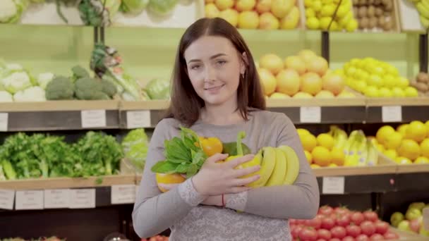 Retrato de la encantadora chica morena posando con fuits en la tienda de comestibles. Joven mujer caucásica sosteniendo plátanos, pomelo y vegetación y sonriendo a la cámara. Estilo de vida vegano, alimentación saludable, comida orgánica . — Vídeos de Stock