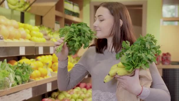 Positive junge Frau riecht Bündel Petersilie im Supermarkt. Porträt eines fröhlichen brünetten kaukasischen Mädchens, das sich für Grün im Lebensmittelgeschäft entscheidet. Veganismus, vegane Lebensweise, gesunde Ernährung. — Stockvideo