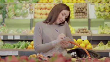 Güzel genç bir bayan telefonda konuşuyor ve süpermarkette notlar alıyor. Marketteki alışveriş listesini netleştiren beyaz kadın gönüllü portresi. İnsanlık, yaşam tarzı, gönüllülük.