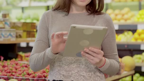 Αγνώριστη νεαρή γυναίκα που ελέγχει λίστα αγορών στο tablet και μαζεύοντας ακτινίδια στο σούπερ μάρκετ. Πορτρέτο της σοβαρής μελαχρινή Καυκάσια κοπέλα αγοράζουν βιολογικά υγιή φρούτα στο παντοπωλείο. — Αρχείο Βίντεο
