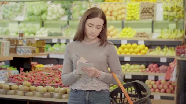 Mujer joven concentrada tomando notas en el supermercado. Retrato de la hermosa morena caucásica chica comprando productos en el supermercado con la lista de compras . — Vídeos de Stock