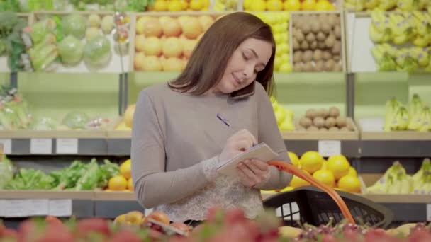 Hübsche junge Frau telefoniert und macht sich im Supermarkt Notizen. Porträt einer charmanten kaukasischen Freiwilligen, die Einkaufsliste im Lebensmittelgeschäft klärt. Menschlichkeit, Lebensstil, Freiwilligenarbeit. — Stockvideo