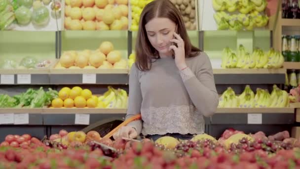 Porträtt av söt ung kvinna med shoppingkorg välja frukt i mataffären och prata i telefon. Brunett säker kaukasiska flicka välja mat i snabbköpet. — Stockvideo