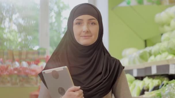 Retrato de una encantadora mujer musulmana posando en el supermercado con una tableta. Joven chica hermosa en hijab mirando a la cámara y sonriendo como de pie en el supermercado. Belleza, estilo de vida, alimentación saludable . — Vídeo de stock