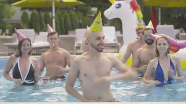 Retrato do jovem homem sorridente em chapéu de festa dançando na piscina de água com pessoas multiétnicas repetindo movimentos no fundo. Animador caucasiano alegre positivo entretendo os hóspedes no resort . — Vídeo de Stock