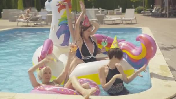Vier fröhliche junge kaukasische Frauen tanzen im Wasserbecken auf Einhorn- und Donut-Schwimmringen. Porträt glücklicher junger schöner Freunde, die an einem Sommertag im luxuriösen Resort ihren Urlaub genießen. — Stockvideo