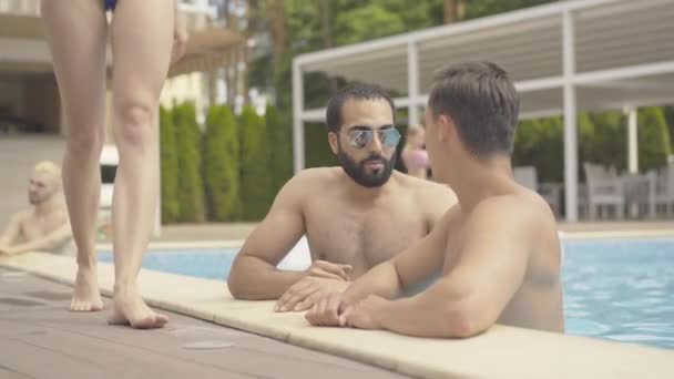Tanımlanamayan zayıf, şehvetli kadın su havuzunda konuşan iki adamın yanından geçiyor. Orta Doğulu ve Kafkasyalı erkekler tatil köyündeki genç kızın ince bacaklarına hayran.. — Stok video