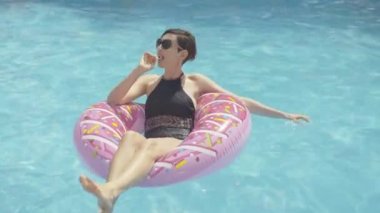 Genç zengin esmer kadın donut yüzme ringinde oturuyor ve lolipop emiyor. Güneşli bir yaz gününde lüks bir tatil köyünde havuzda dinlenen kendine güvenen beyaz bir kadın..